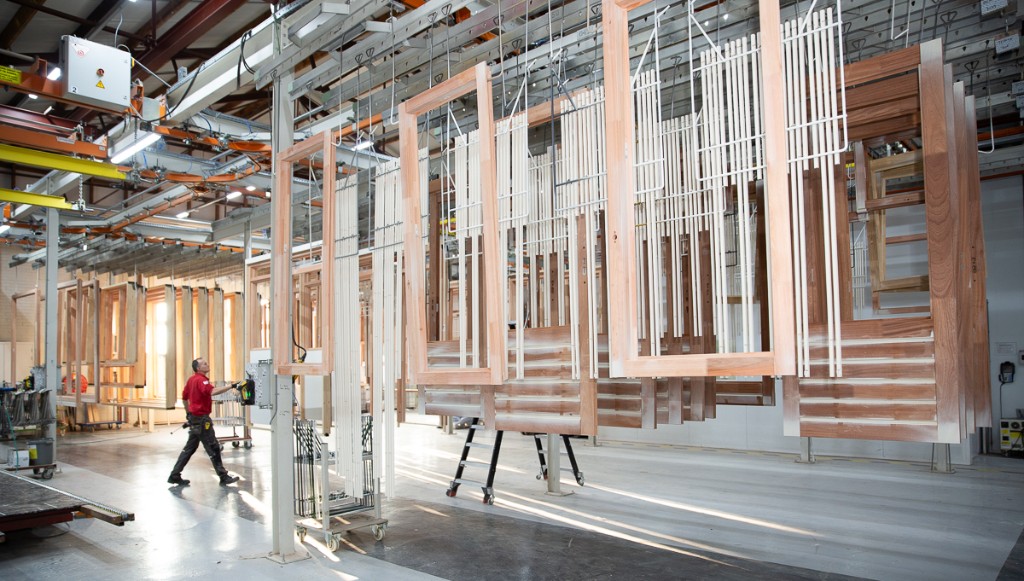 Worker on the production floor walking past wooden window frames hanging on an overhead conveyor